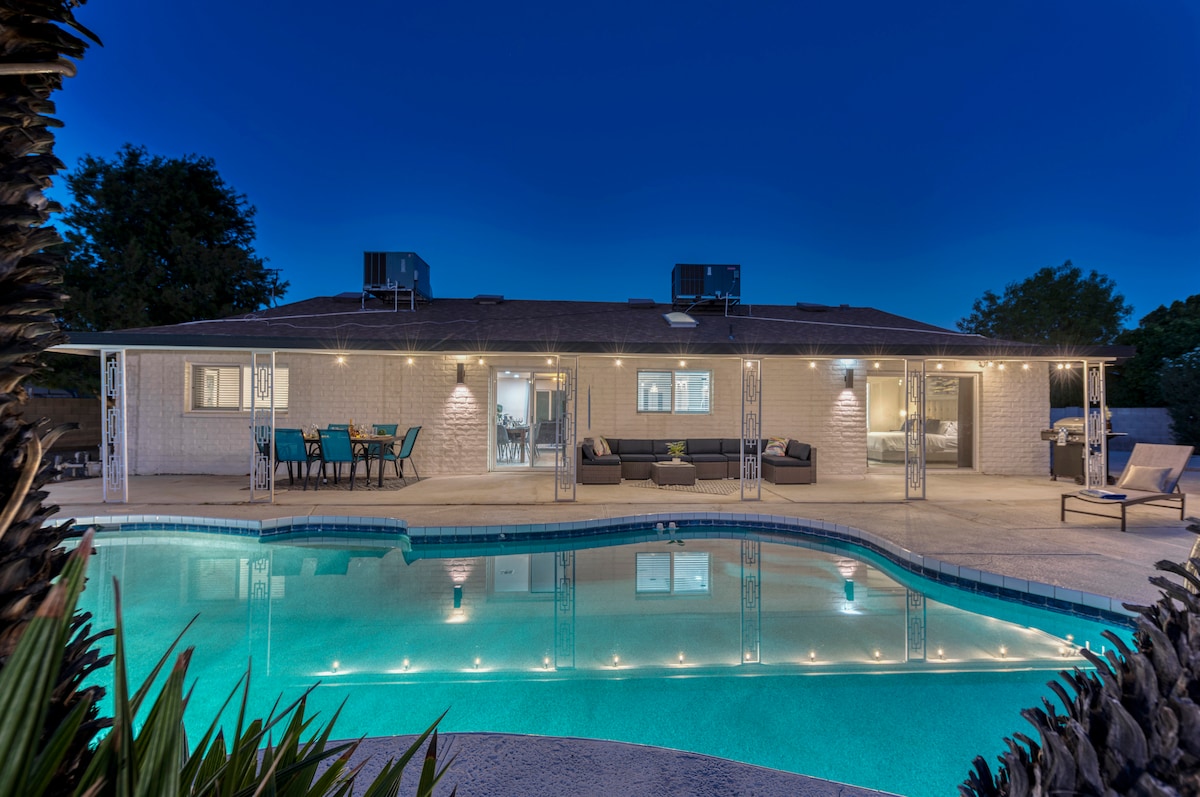 A spacious backyard features a heated swimming pool, surrounded by comfortable lounge seating and a dining area. The home is positioned to create a welcoming atmosphere, with outdoor lighting illuminating the area, viewed against the evening sky.