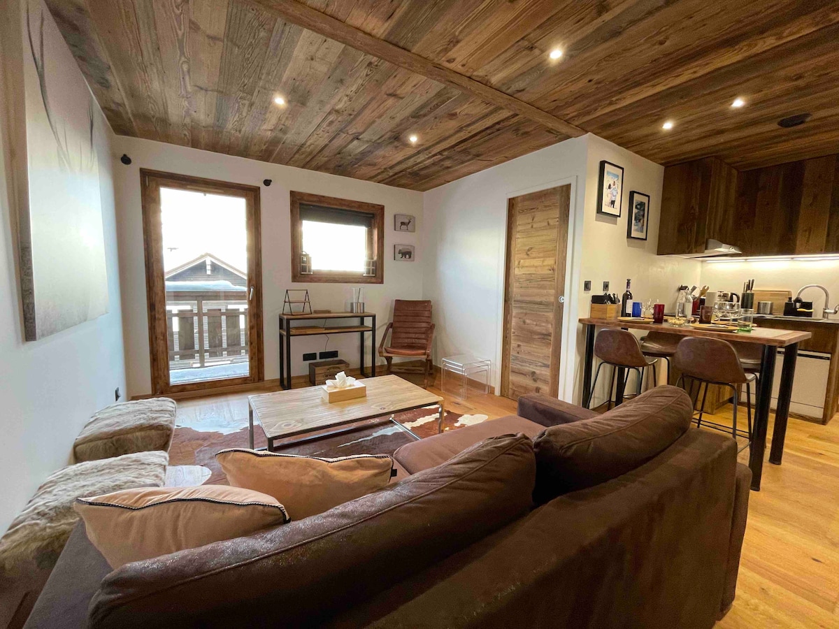 A cozy living area is showcased with wooden beams and a warm color palette. A comfortable sofa is positioned next to a coffee table, with sunlight filtering through large doors that open to a balcony. A dining area and kitchen, featuring modern appliances, are visible in the background.