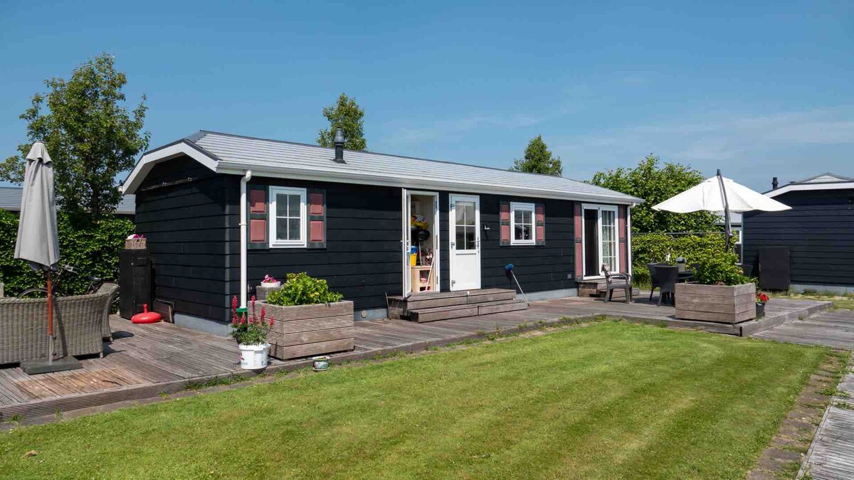 A cozy vacation home is situated in a green setting, with a dark wood exterior and light-colored trim. The decking provides an outdoor space for relaxation, featuring seating arrangements and planters. Umbrellas offer shade, and the surrounding lawn contributes to a serene atmosphere.