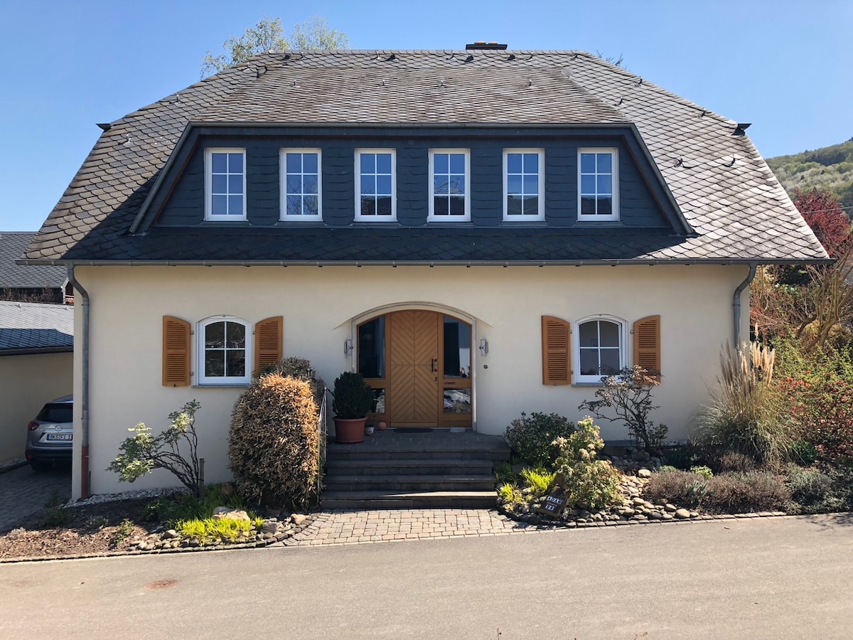 A charming two-story house features a neutral facade complemented by wooden shutters. A welcoming entrance with a wooden door is framed by greenery and flowering plants. Symmetrical windows enhance the building's appeal, and a stone pathway leads up to the entrance.