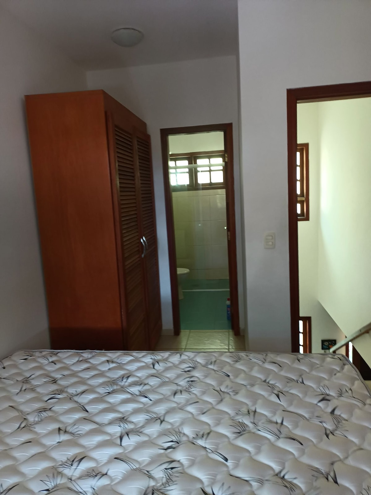 A private room features a spacious bed with a partially visible mattress. Wooden cabinets occupy one side, and an open doorway leads to a bathroom area, with light filtering in through small windows.