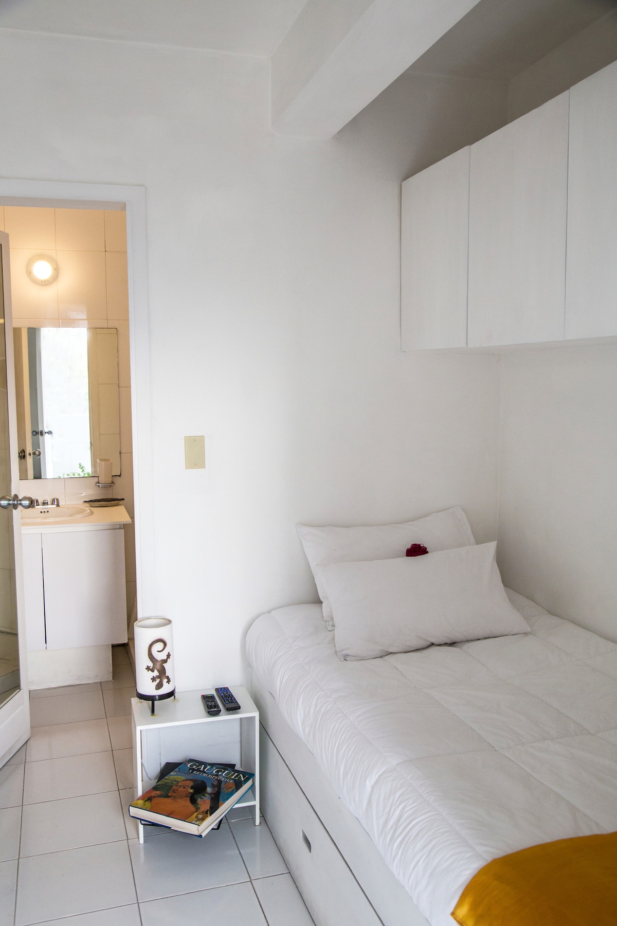 A small room features a single bed with a white bedspread and decorative cushions. A compact nightstand holds a lamp and remote controls. A doorway leads to a private bathroom equipped with modern fixtures. The space is bright, with light-colored walls and tiled flooring.