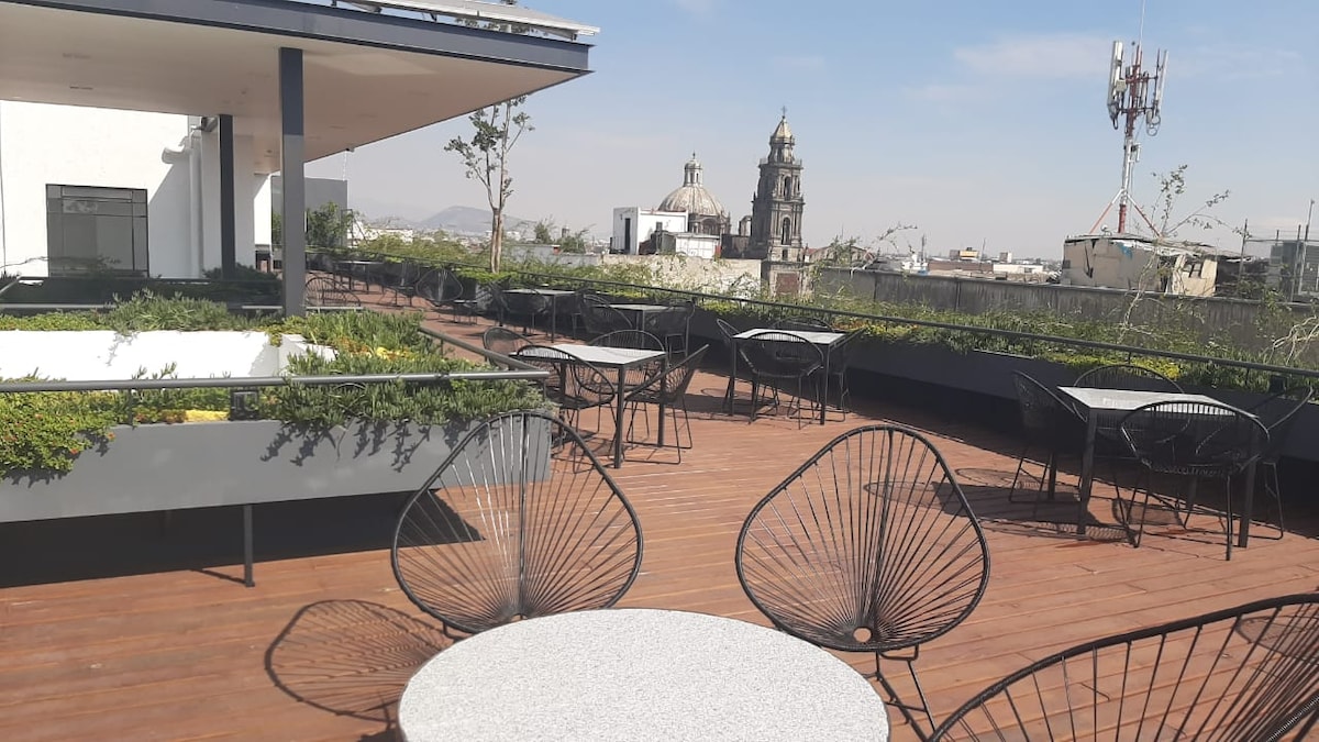 A spacious rooftop area features wooden flooring with several black metal chairs and tables arranged thoughtfully. Greenery decorates the edges of the terrace, and a historic building's dome is visible in the background against a clear sky.