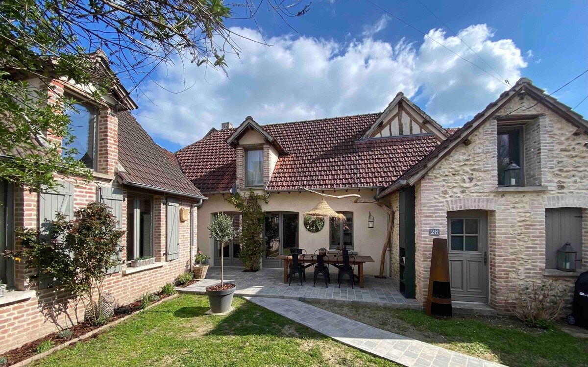 The exterior of the charming house features a mix of brick and stone façades. A dining table with chairs is situated under a canopy in the garden, surrounded by well-maintained greenery. The sky above is partly cloudy, creating an inviting atmosphere.