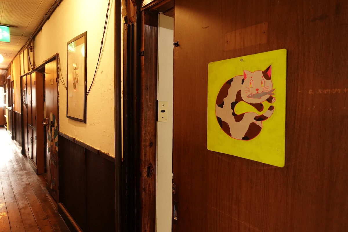 A hallway with wooden flooring is visible, featuring a door adorned with a colorful cat painting. Framed artwork decorates the walls, adding character to the space. Soft, warm lighting enhances the welcoming ambiance.