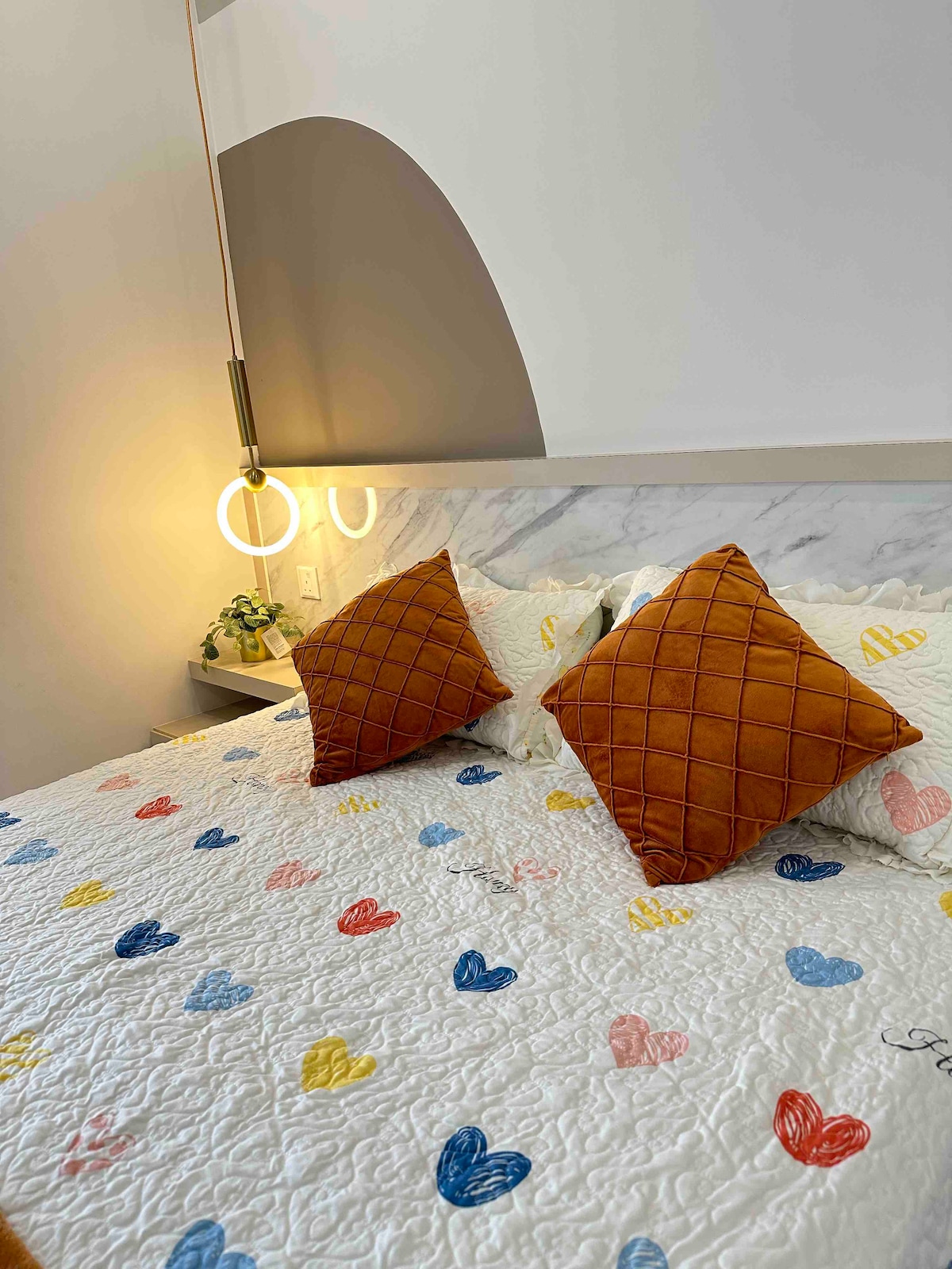 A spacious bed is adorned with a white quilt featuring colorful heart patterns. Two decorative cushions in a chocolate brown tone are placed at the head of the bed. Soft lighting from a nearby bedside lamp offers a gentle glow, complementing the cozy ambiance.