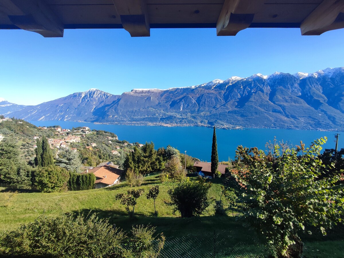 A panoramic view of the lake and surrounding mountains is framed by the balcony's wooden structure. Lush green hills lead down to the shimmering water, dotted with quaint homes. The clear blue sky enhances the scenic landscape, creating an inviting outdoor atmosphere.