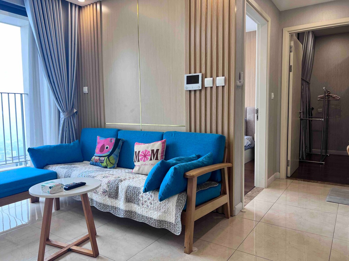 A modern living space features a bright blue sofa adorned with decorative pillows. A small wooden table is positioned in front of the sofa. The walls are light-colored, and large windows allow natural light to brighten the area.