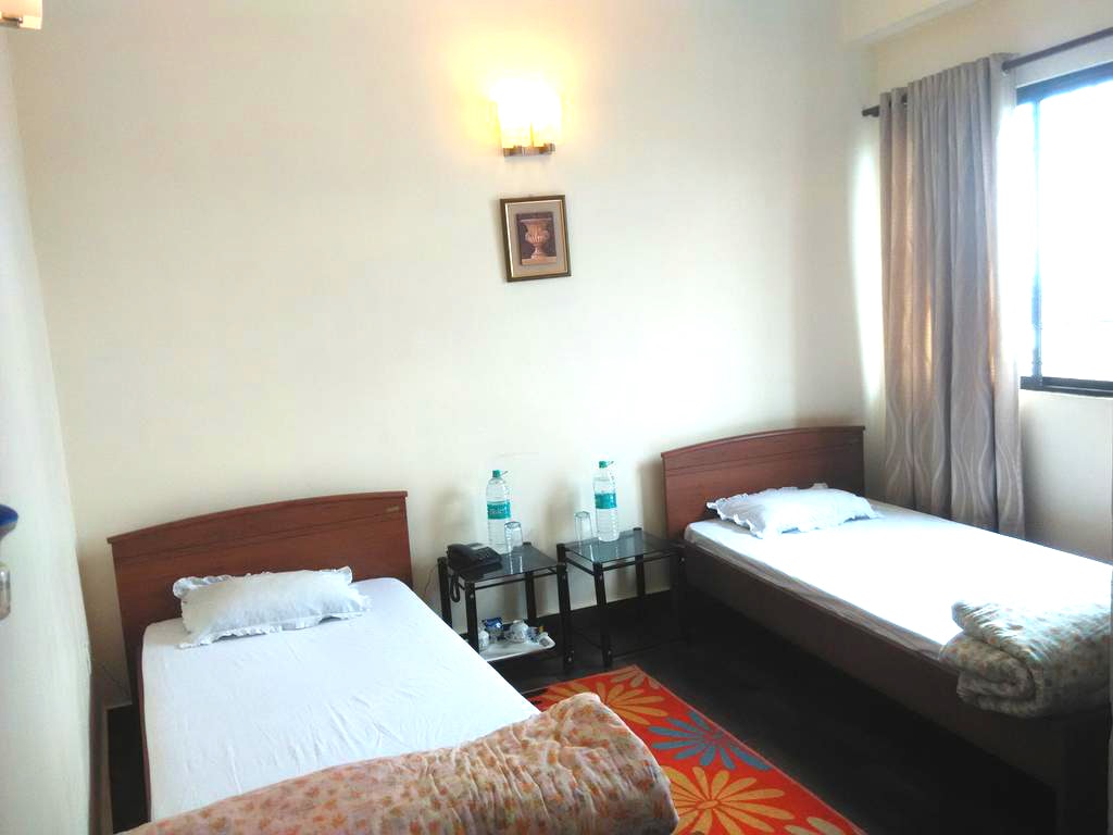 The room features two single beds with light bedding, situated beside a small table. Natural light enters through a window adorned with light curtains. A wall-mounted light fixture and a framed picture add to the simple decor. Bottled water is placed on the table.