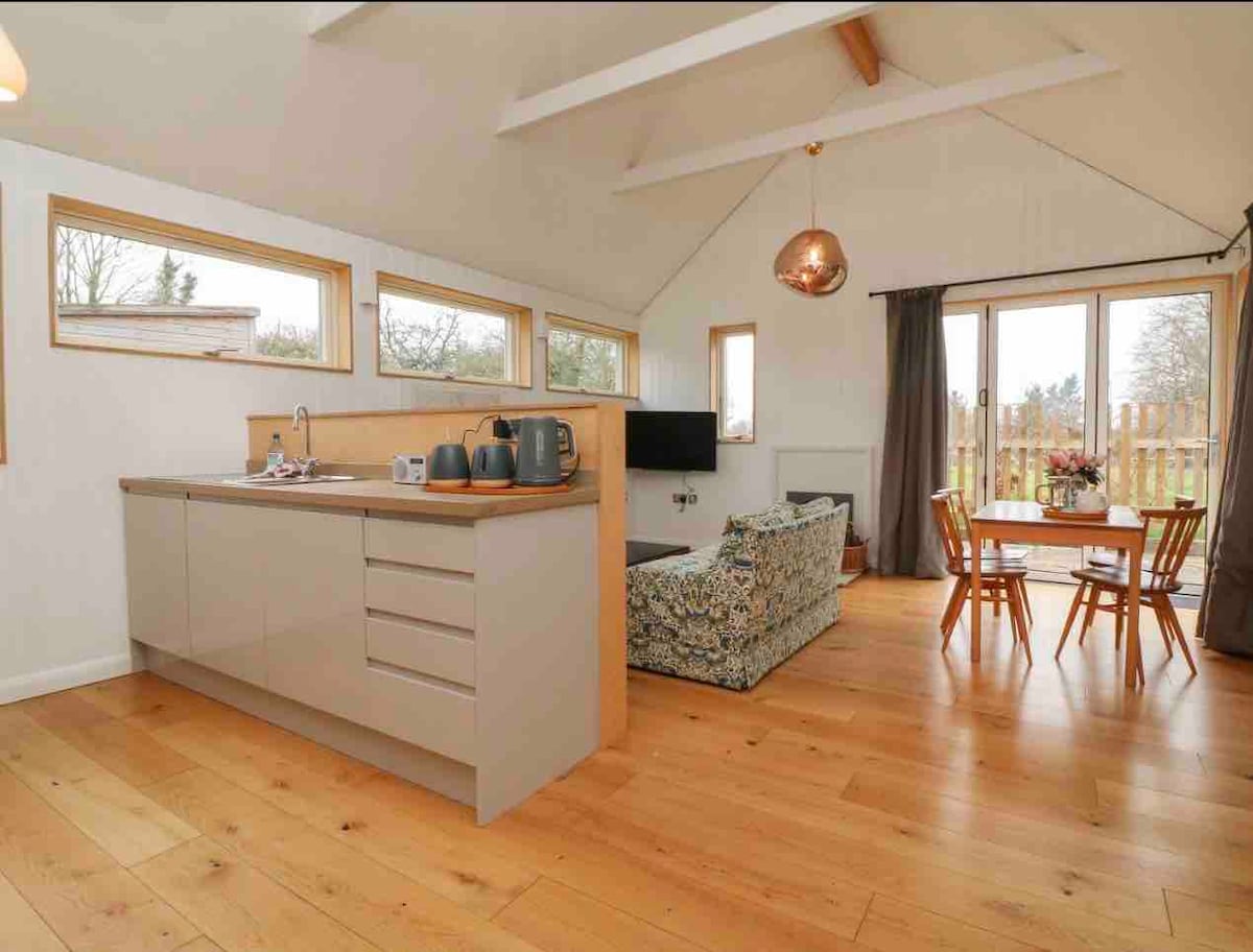 An open-plan living area is visible, featuring a modern kitchen with sleek cabinetry and kitchenware. Natural light streams through large windows, illuminating the cozy seating area and dining table. Warm wooden flooring complements the inviting atmosphere, enhancing the interconnected spaces.
