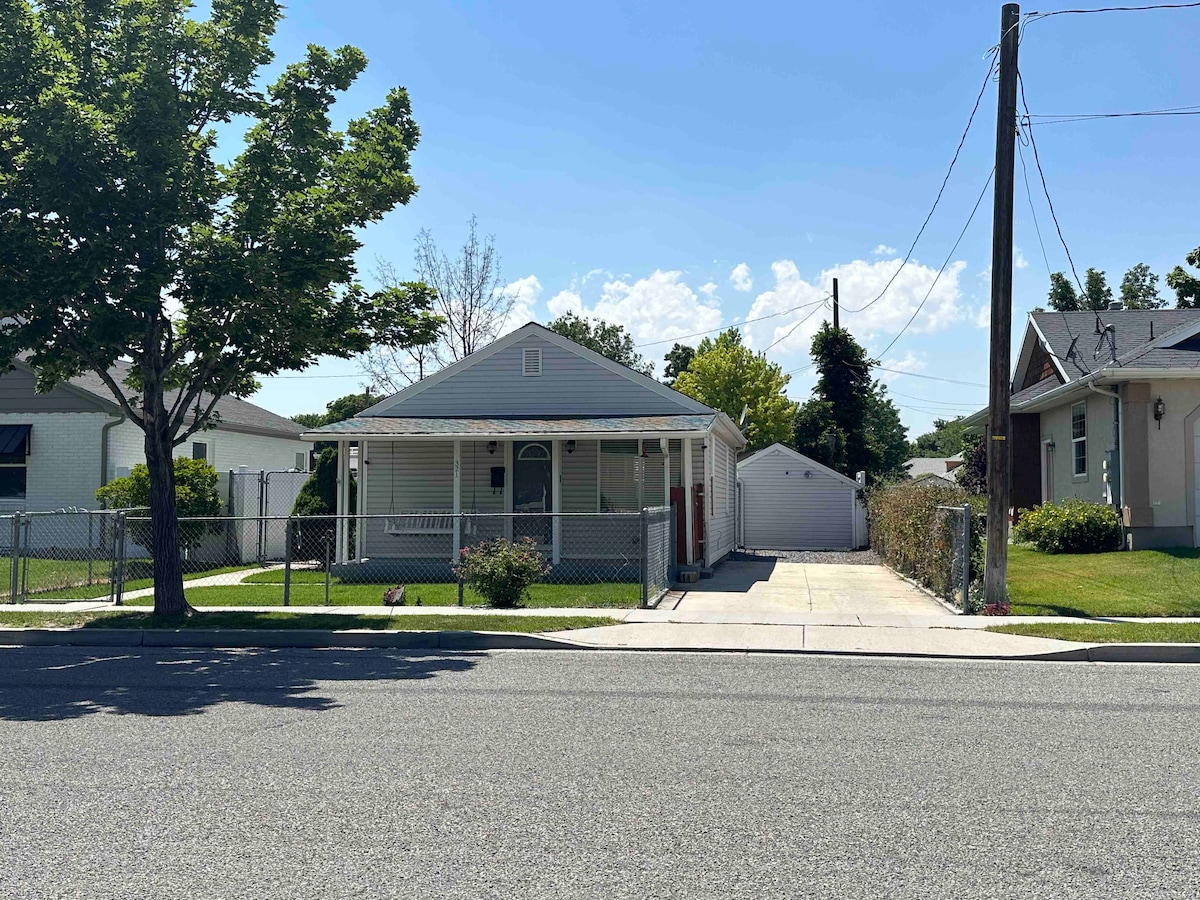 The exterior of the single-story cottage is framed by a well-maintained fence and mature trees. A spacious front porch is visible, providing seating options. A driveway leads to a detached garage, and the surrounding area includes grass lawns and additional houses.