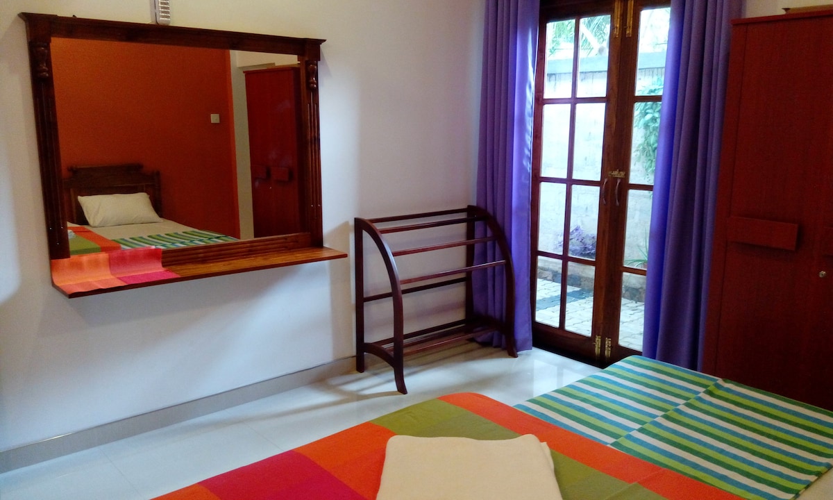 A bright sleeping area features a spacious bed with a colorful striped blanket. A mirror reflects the vibrant orange and purple walls. A wooden rack stands nearby, while large glass doors open to a small outdoor space, allowing natural light to enter the room.