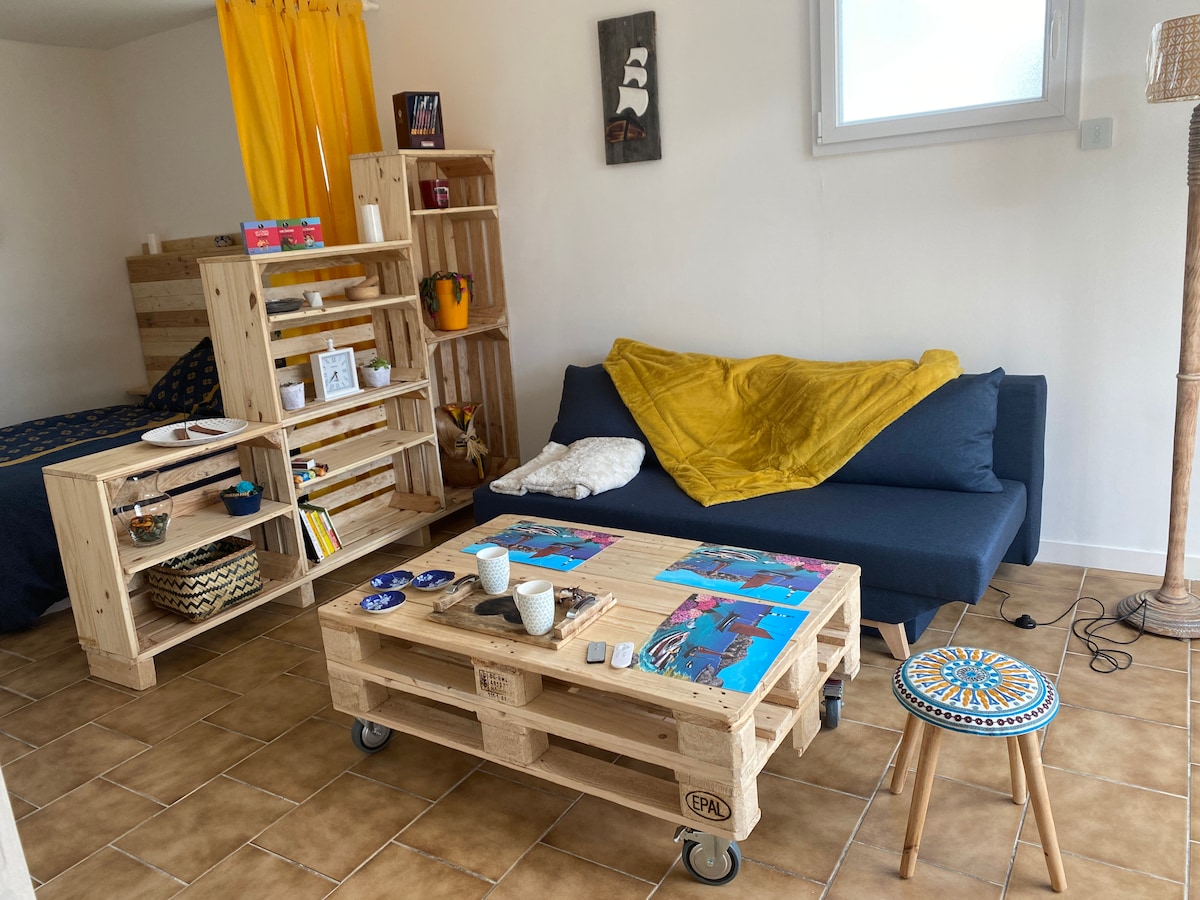 A cozy living area features a blue sofa adorned with a yellow throw. A wooden coffee table with colorful placemats sits in front of the sofa. Shelving units display decorative items, and a small stool completes the space, which is illuminated by natural light.