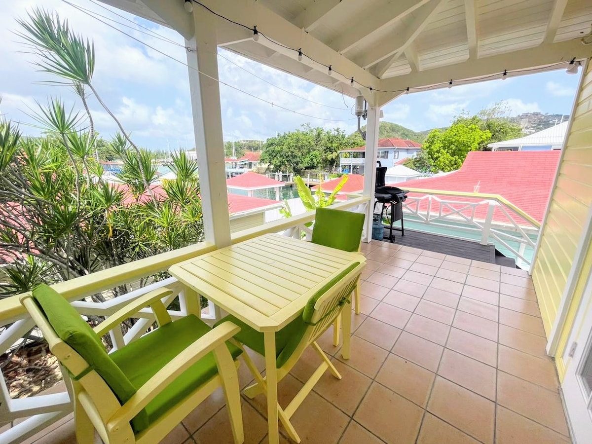 A covered balcony is shown with a light wood table and two green chairs positioned for dining. Lush greenery is visible nearby, along with a barbecue grill. The open space offers glimpses of the harbor and surrounding colorful buildings.