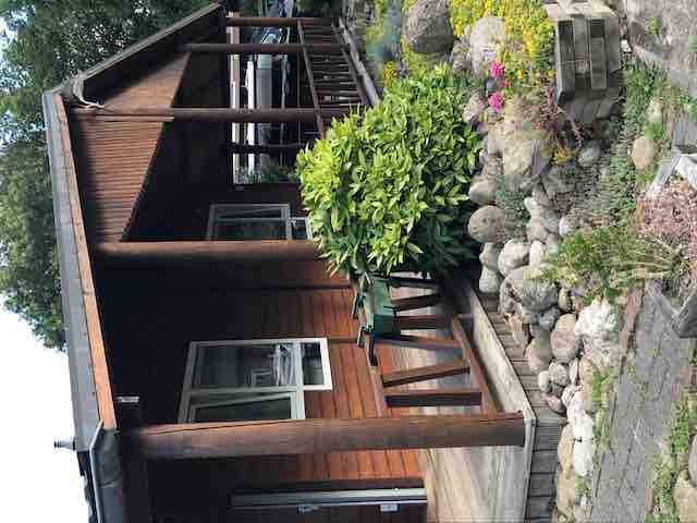 The exterior of the chalet showcases rustic wooden elements, complemented by a covered porch. Lush greenery and varied plant life surround the entrance, with a pathway leading towards the stone garden. Large windows provide natural light, enhancing the inviting ambiance of the home.