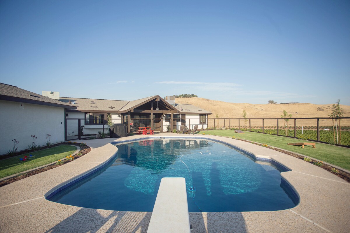 The exterior features a large, inviting pool set within a spacious yard, surrounded by fencing and greenery. A shaded patio area with wooden beams overlooks the pool, while the hillside in the background adds to the serene atmosphere.
