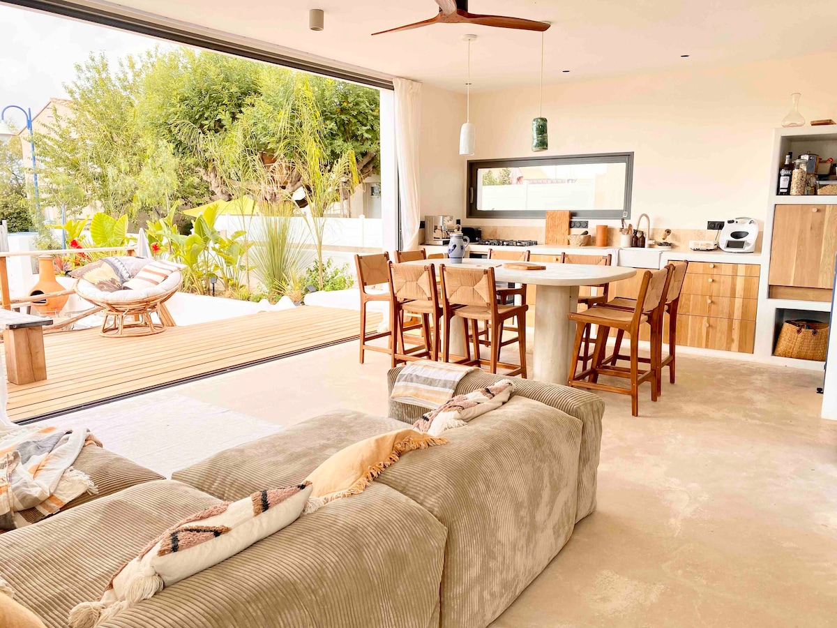 An open living area features a spacious sofa and a circular dining table surrounded by wooden chairs. Large glass doors slide open to reveal a wooden deck adorned with tropical plants, creating an inviting connection between indoor and outdoor spaces.
