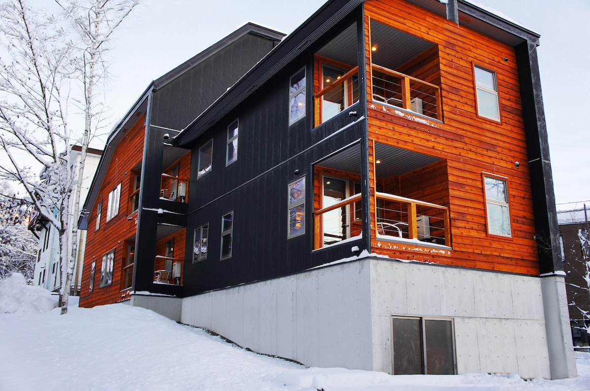 The image showcases the exterior of a newly constructed three-story building. The lower portion features a concrete base, while the upper levels are adorned with contrasting dark timber and bright orange cladding. Balconies with wooden railings are visible on each floor, creating a modern aesthetic.