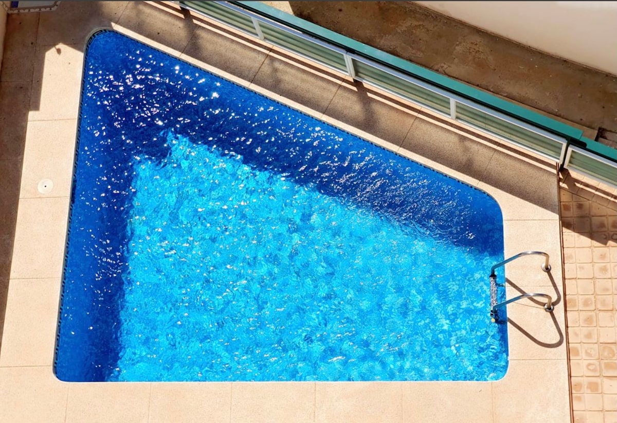 The image features a triangular-shaped pool, filled with clear blue water that reflects sunlight. A metal ladder is positioned at one end, providing access to the pool, while the surrounding area is paved with light-colored tiles.