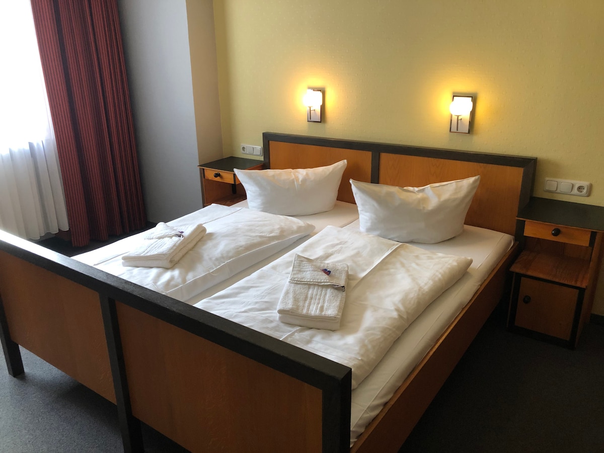 A well-appointed sleeping area is furnished with a spacious bed dressed in white linens and topped with two pillows. Soft lighting is provided by wall-mounted lamps above the bed, while a window dressed with light curtains allows natural light to enter the room.