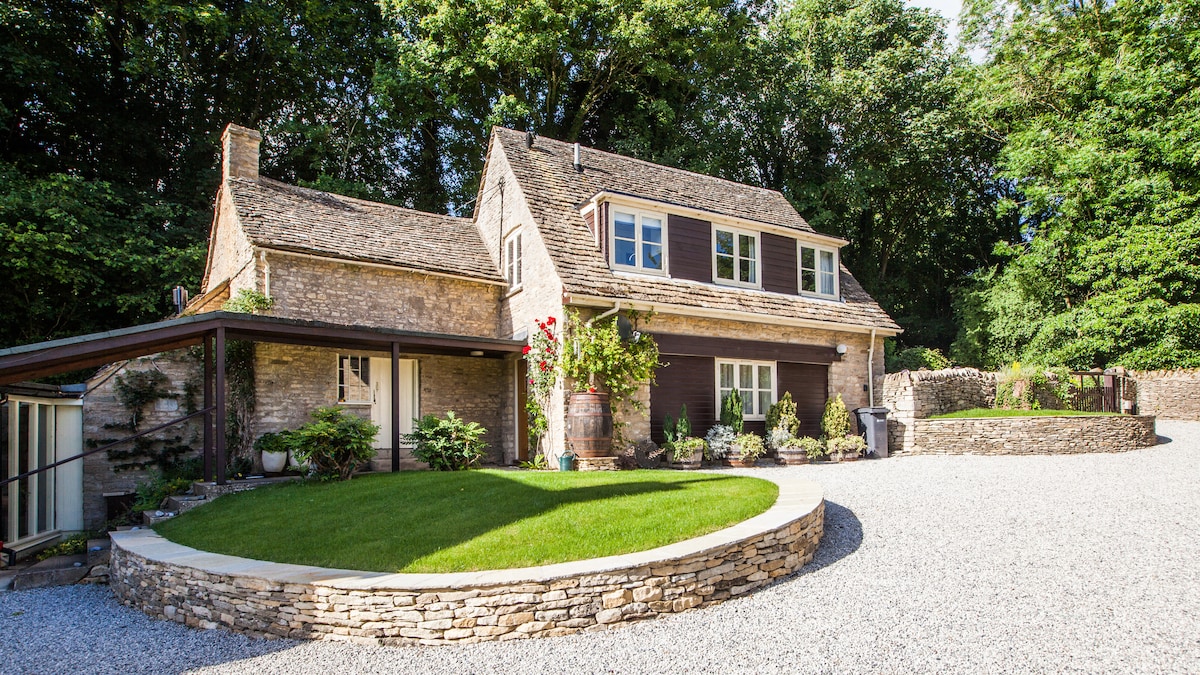 A charming stone-built lodge is set within a lush green landscape. The private entrance leads to a circular lawn surrounded by a gravel driveway and flower pots. Tall trees provide a natural backdrop, enhancing the sense of seclusion and tranquility.