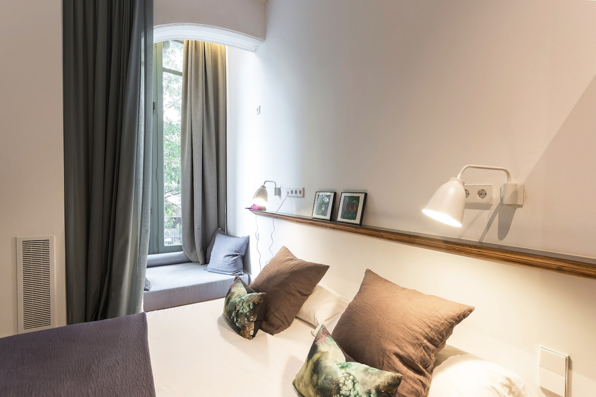 A bright sleeping area is featured, showcasing a spacious bed adorned with decorative pillows. Two bedside lamps illuminate the space. A corner window with soft curtains offers a view of the outside, while a small seating area with cushions adds to the comfort.