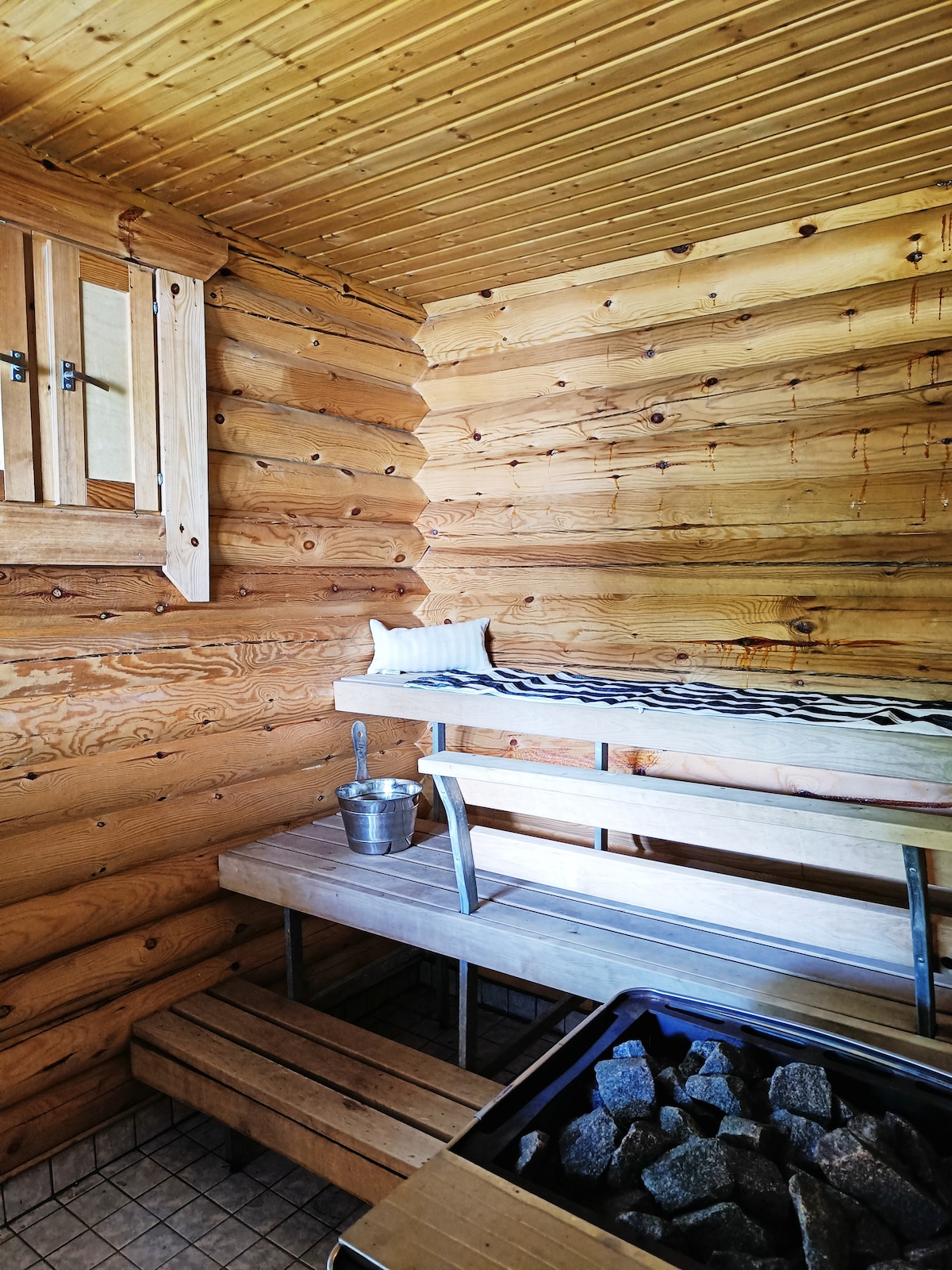 Inside the sauna, wooden walls create a warm, natural atmosphere. A bench is positioned against the wall, topped with a small cushion. An area holds a bucket and ladle, with a stove featuring stones below, completing the traditional sauna experience.