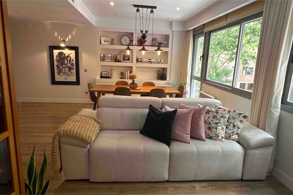 A spacious living area features a comfortable light-colored sofa with decorative cushions. A wooden dining table is positioned in the background, accompanied by several chairs. Large windows allow natural light to illuminate the space, offering a view of greenery outside.