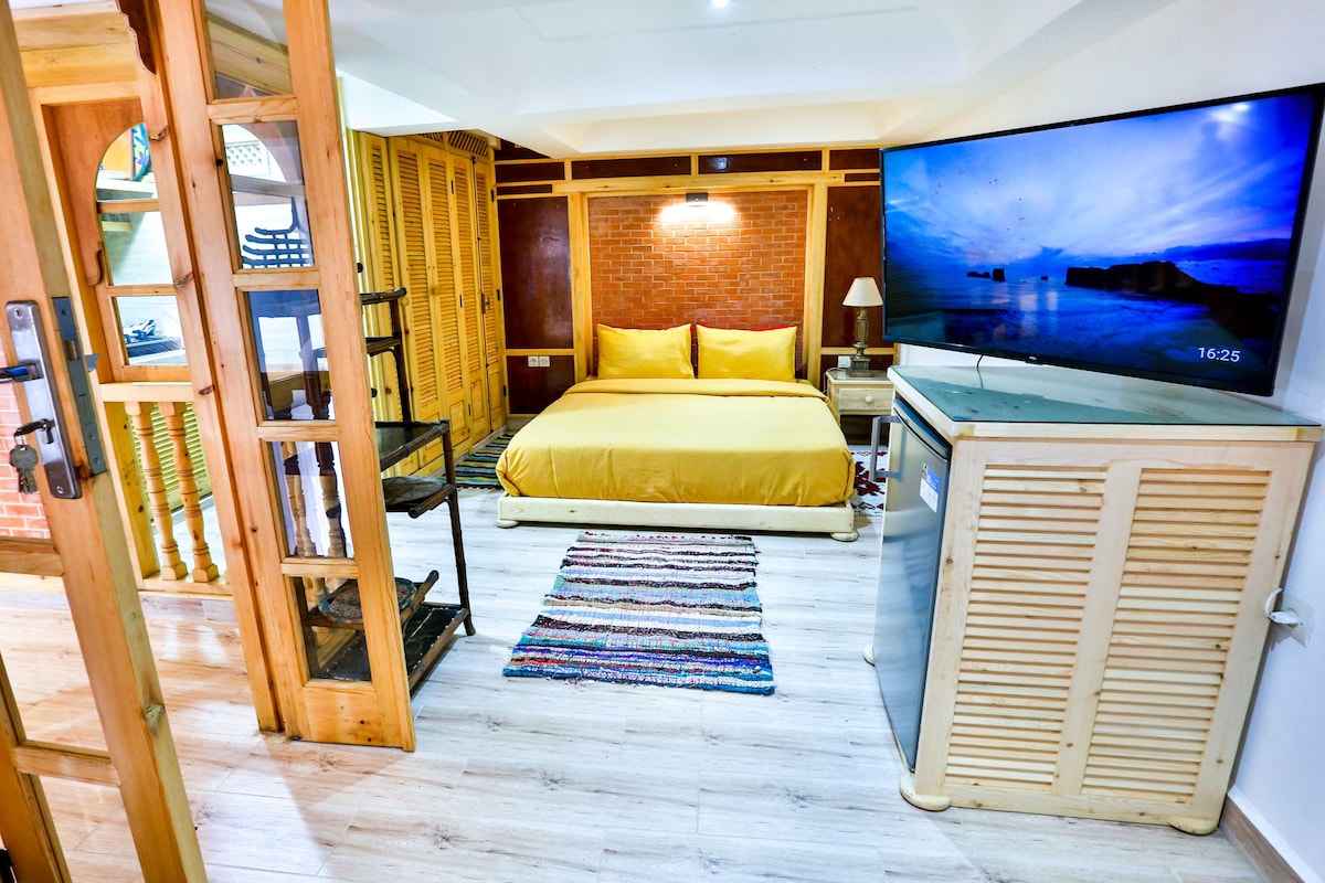A cozy sleeping area features a spacious bed with warm yellow bedding against a brick accent wall. A large screen television is mounted on the wall. Light wood cabinetry and a small refrigerator are present, enhancing the practical charm of the space.