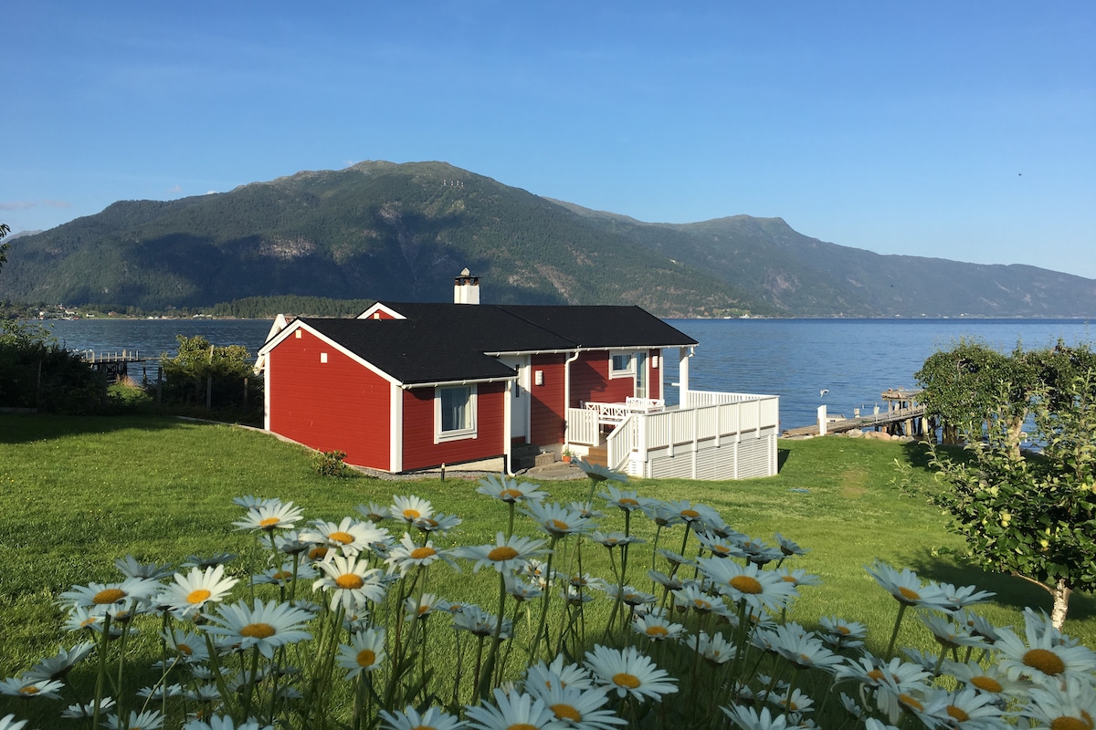 The red cottage is positioned by the seashore, framed by lush green grass and blooming daisies in the foreground. Majestic mountains rise in the background under a clear blue sky, providing a serene and natural setting.