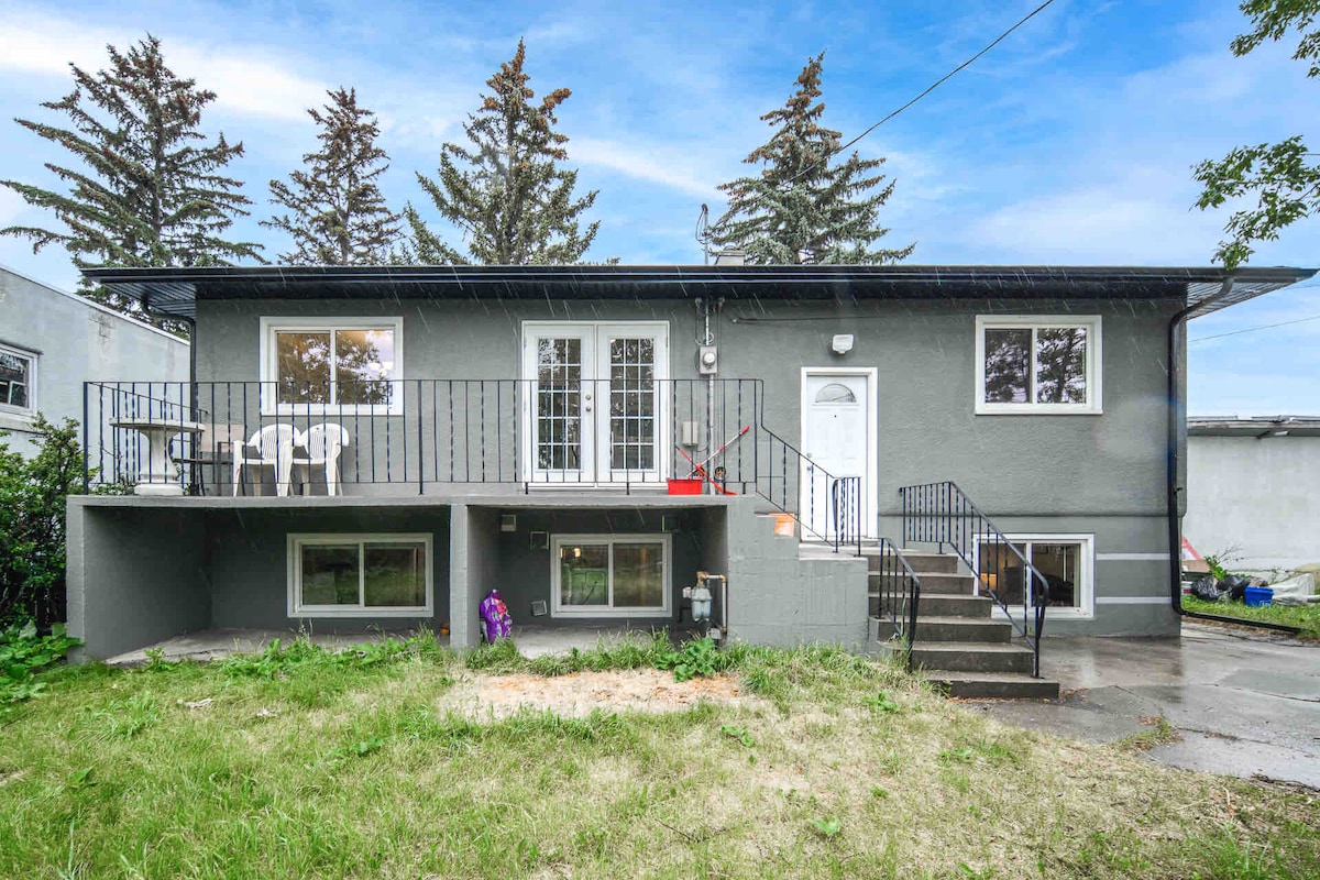 The exterior of the bungalow showcases a two-level design with a grey facade. A staircase leads to the upper entrance, flanked by a small patio with four white chairs. Large windows are visible on both levels, surrounded by green grass and pine trees.