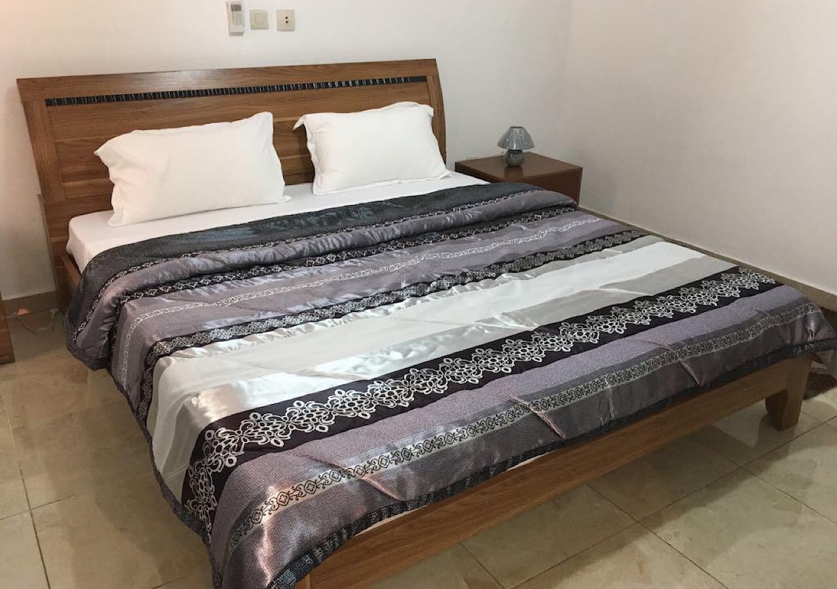 A spacious bed with a decorative patterned blanket is centered in the room. Two white pillows rest at the head of the bed. A wooden nightstand sits adjacent, featuring a bedside lamp. Neutral-toned tiled flooring complements the overall aesthetic of the space.