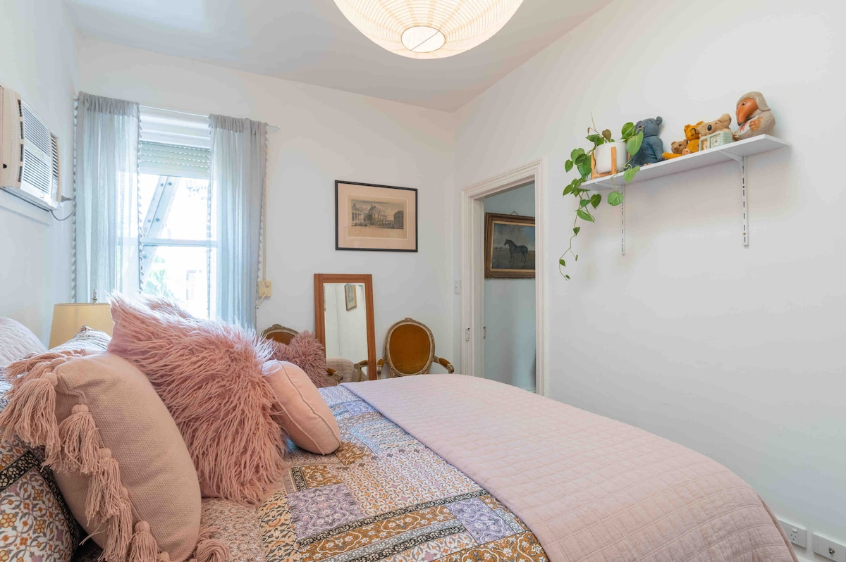 A cozy bedroom features a spacious bed adorned with decorative pillows and a soft blanket. A vintage mirror and plush seating are visible, while a shelf displays potted plants and small figurines. Natural light enters through a window, enhancing the room's inviting ambiance.