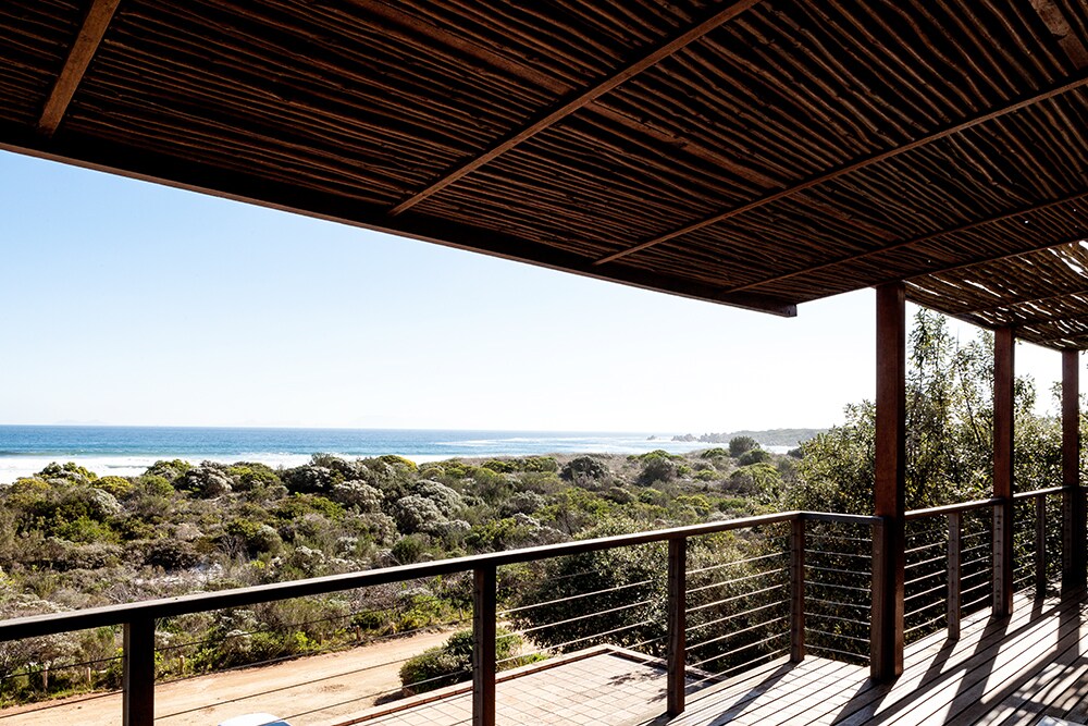 A spacious balcony is covered with natural wood finishes, offering a panoramic view of the ocean and surrounding greenery. The railing showcases sweeping vistas of the beach and coastline, with a clear blue sky above. Natural light highlights the serene atmosphere.