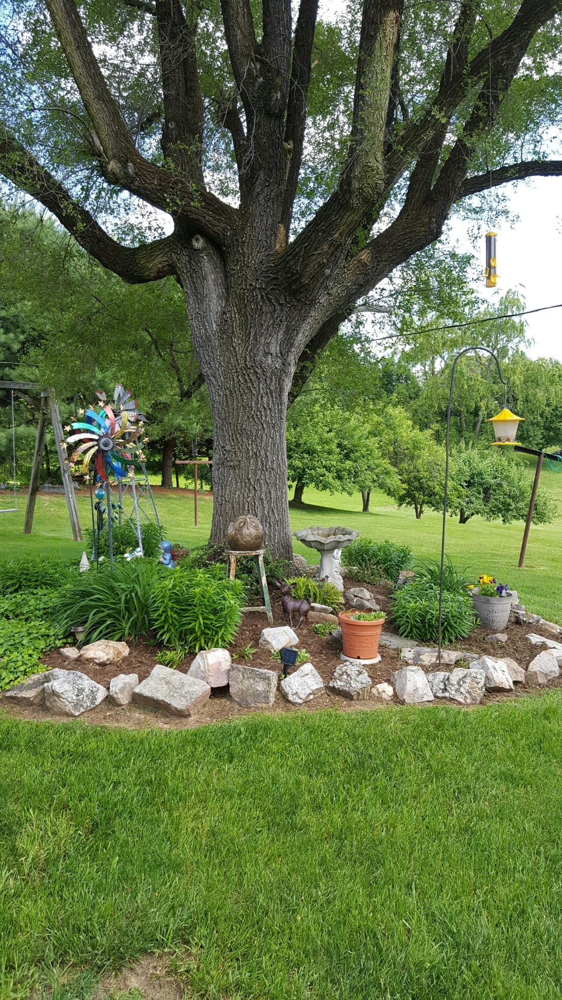 A large tree stands in a lush green backyard, surrounding a decorative garden area. Colorful wind spinners and a bird feeder are visible, while rocks and plants create defined borders. A birdbath and potted plants add charm to this inviting outdoor space.