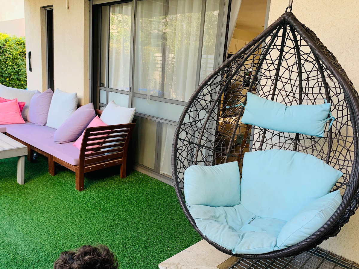A cozy outdoor seating area features a hanging chair with soft blue cushions and a variety of decorative pillows on a wooden bench. Lush green grass creates a comfortable and inviting setting. Large glass doors provide access to the indoor living space.