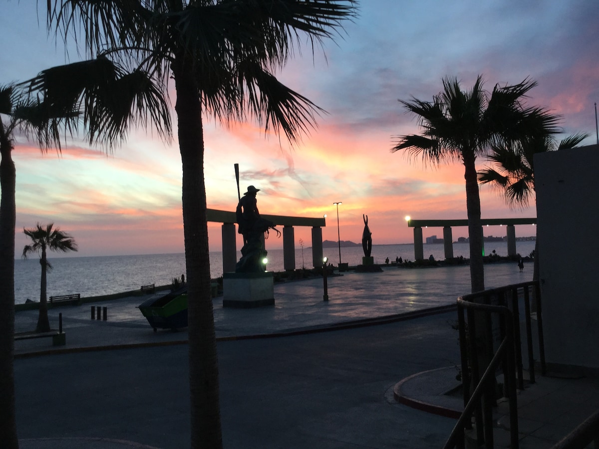 A vibrant sunset casts soft hues over the Malecon, framed by palm trees. Silhouetted sculptures stand gracefully along the promenade, with the tranquil Sea of Cortez reflecting the evening colors. Streetlights begin to glow, adding warmth to the serene atmosphere.
