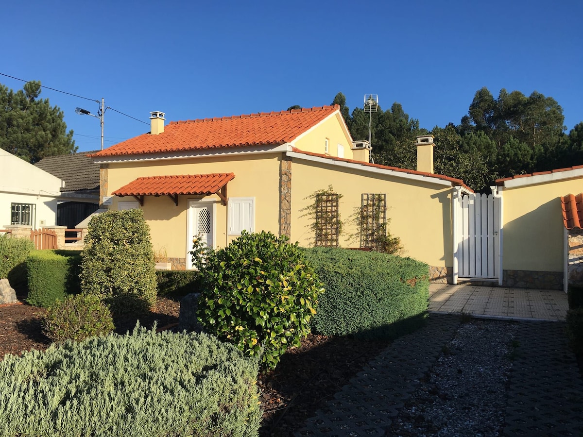 A charming yellow house with a red-tiled roof is surrounded by neatly trimmed shrubs and greenery. The entrance features a white gate, leading to a paved path along a well-maintained garden. Clear skies and sunlight enhance the inviting appeal of the exterior.