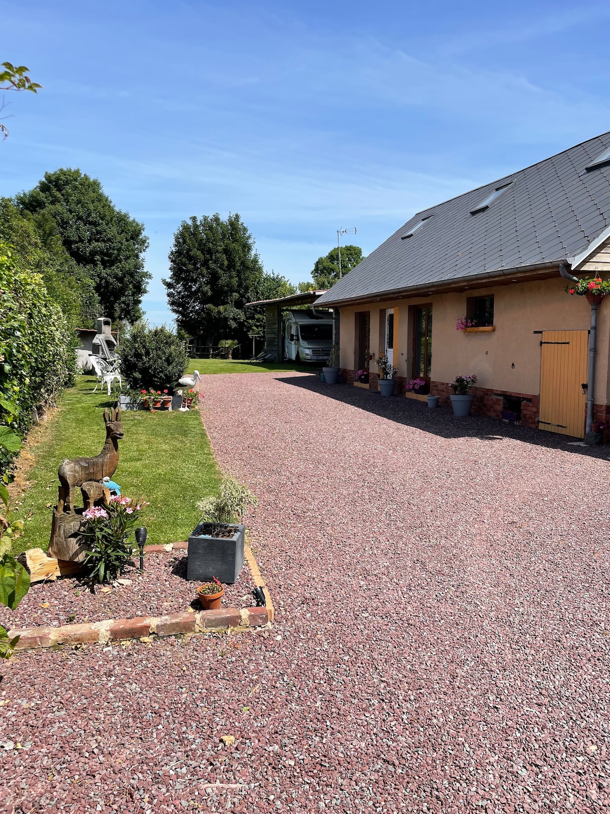A gravel pathway leads to the entrance of a charming gite, flanked by neatly arranged flower pots and greenery. A small decorative statue is positioned among the plants, while the building features a warm-toned exterior and a sloped gray roof under a clear blue sky.