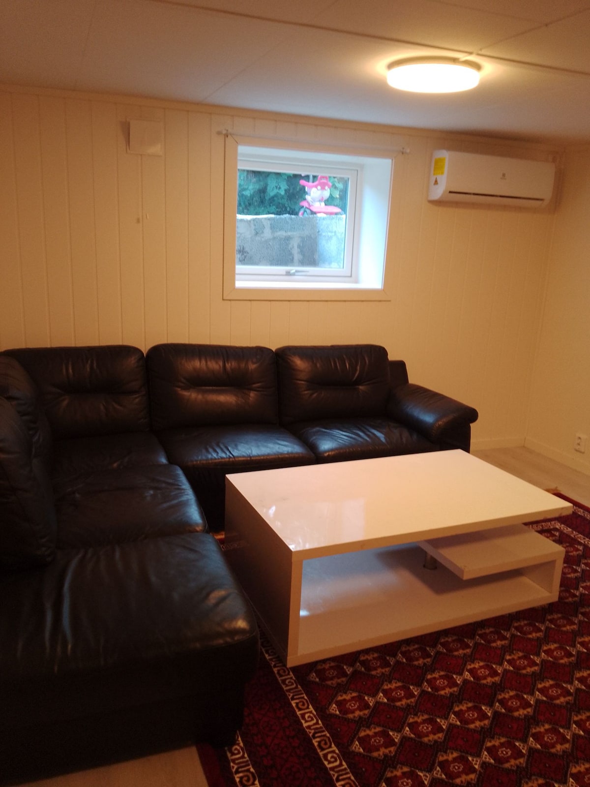 A spacious living area features a black leather sectional sofa arranged around a white coffee table. Natural light enters through a window, brightening the room's neutral-toned walls. A decorative rug adds warmth to the floor.