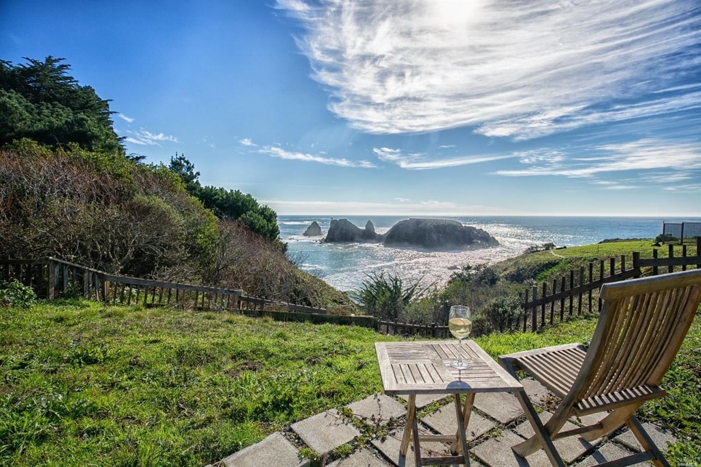 A wooden lounge chair and side table are positioned on a patio, offering a clear view of the ocean and rocky coastline. Lush greenery and low bushes outline the property, while a bright blue sky with wispy clouds complements the serene setting.