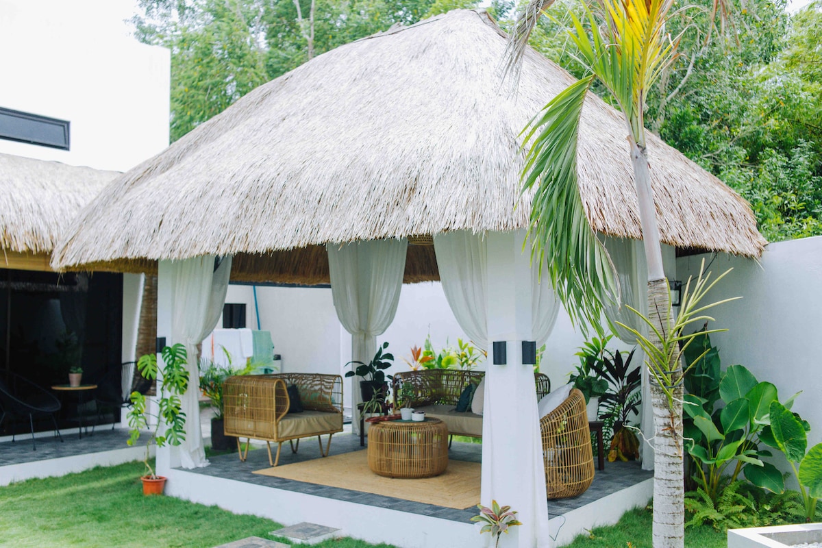 A spacious gazebo is adorned with a thatched roof, providing shade and comfort. Comfortable seating arrangements are complemented by plants and greenery, creating a tranquil outdoor lounge area. Soft drapes frame the space, enhancing the inviting atmosphere for relaxation.