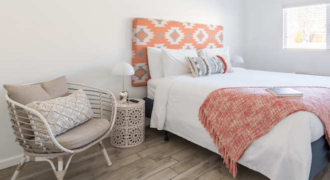 A spacious bedroom features a large bed with a colorful patterned headboard. A cozy throw blanket is draped across the bed. A decorative side table stands next to a stylish chair, which is accented with cushions. Natural light filters through a window covered by light blinds.