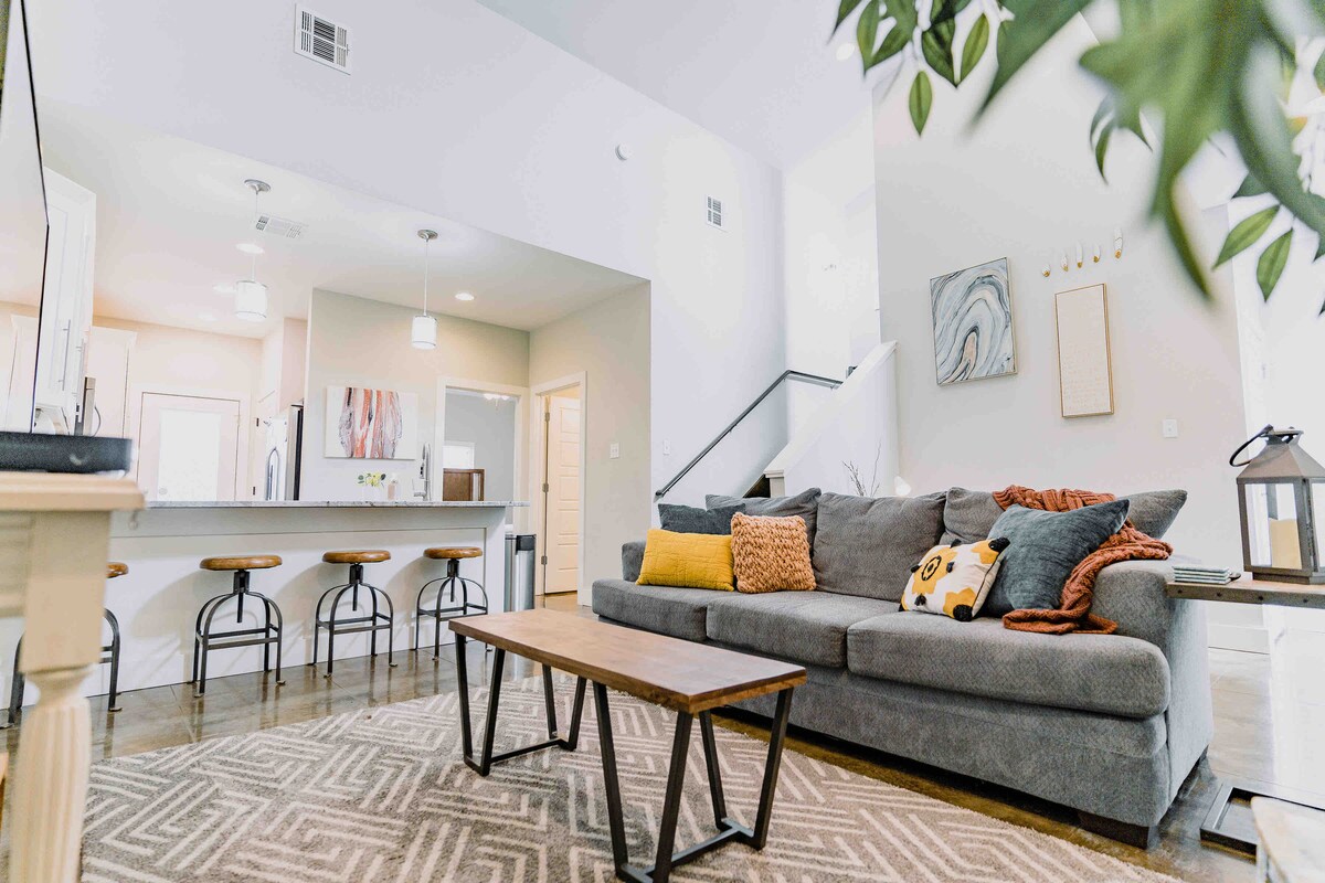A spacious living area is shown, featuring a comfortable sofa adorned with accent pillows and a cozy throw. A large coffee table is centrally placed, while barstools are seen along a kitchen island in the background. Natural light brightens the room through the open layout.