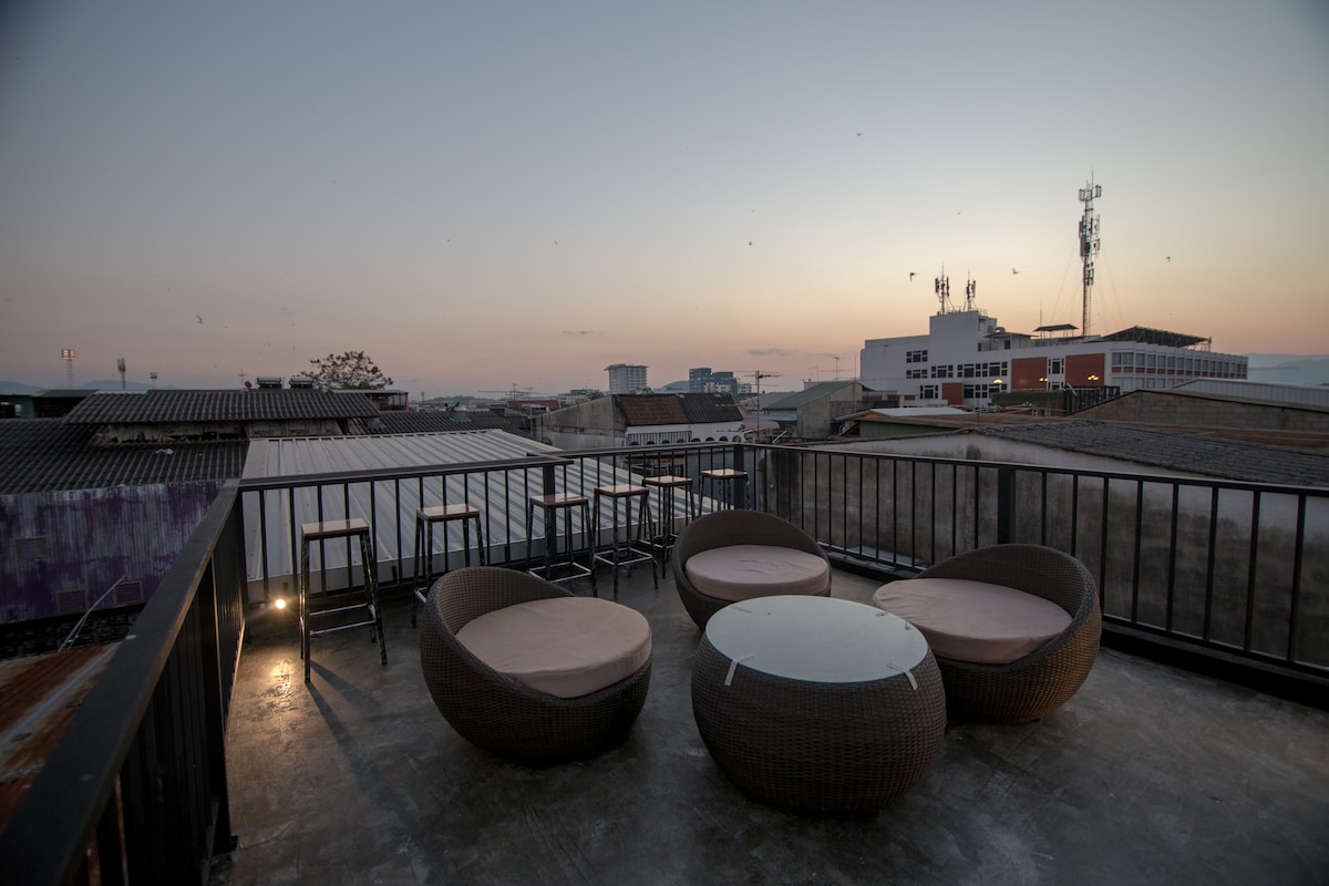 A rooftop terrace offers a series of circular lounge seats surrounding a central table. The scene captures a warm sunset, with city buildings and telecommunication towers visible on the horizon. The design promotes casual gatherings and views of the surrounding area.