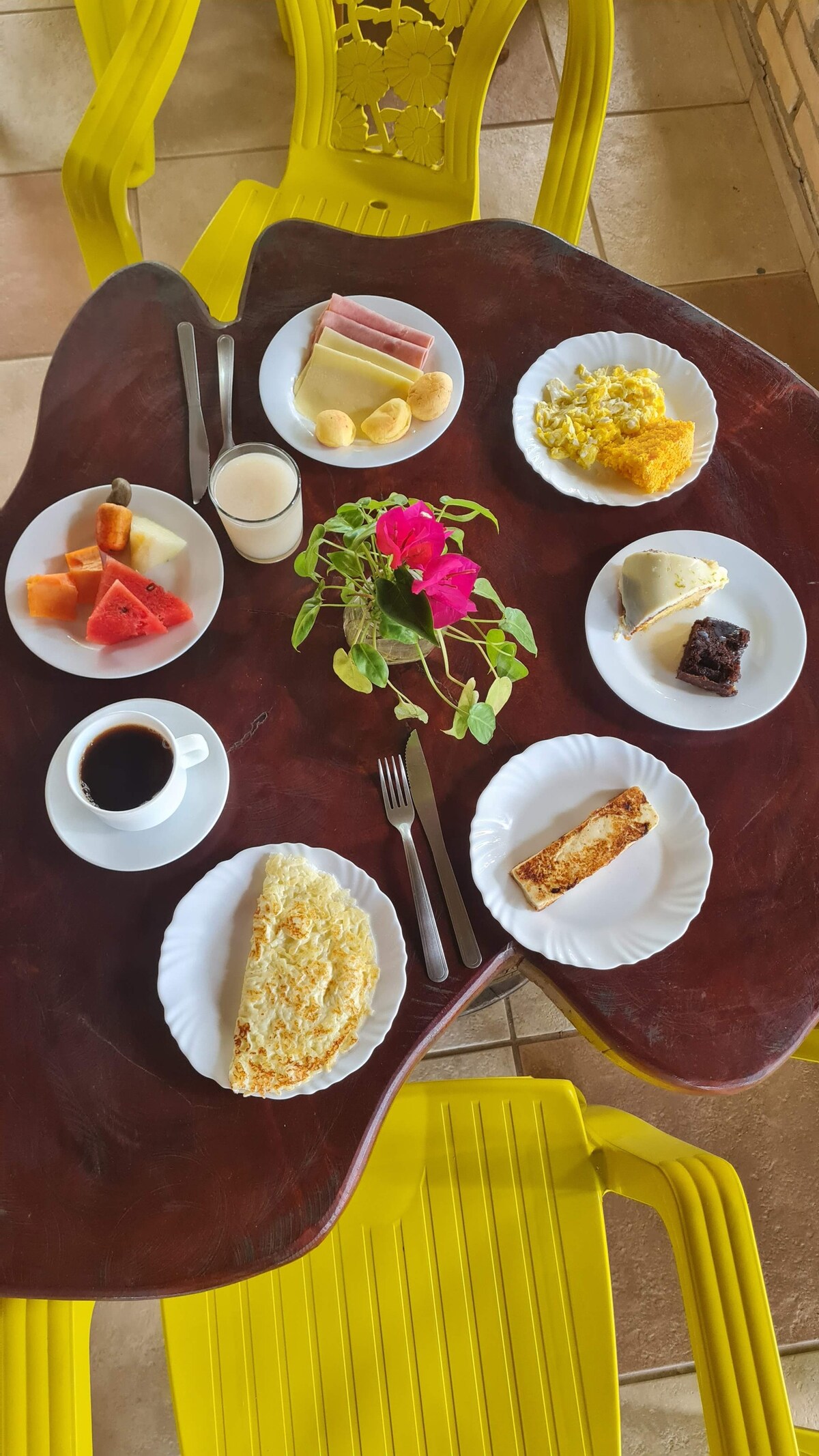 A wooden table is set with white plates featuring a variety of breakfast items, including fruits, eggs, ham, and baked goods. A cup of coffee and a glass of juice are also present. The bright yellow chairs add a cheerful touch to the dining area.