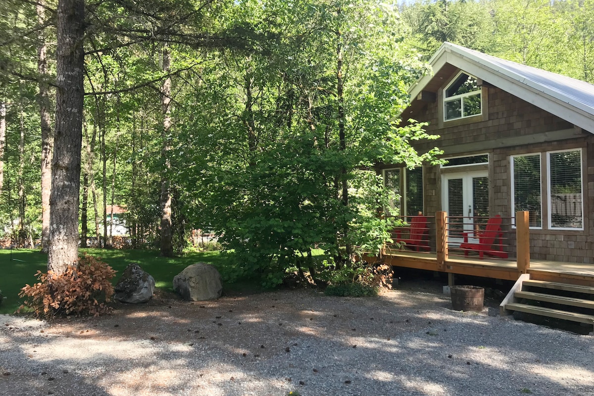 A cabin with a wooden exterior is partially surrounded by greenery, indicating a serene outdoor setting. A prominent deck features two red chairs, creating a welcoming entry. Rocky natural landscaping complements the lush lawn and surrounding trees.