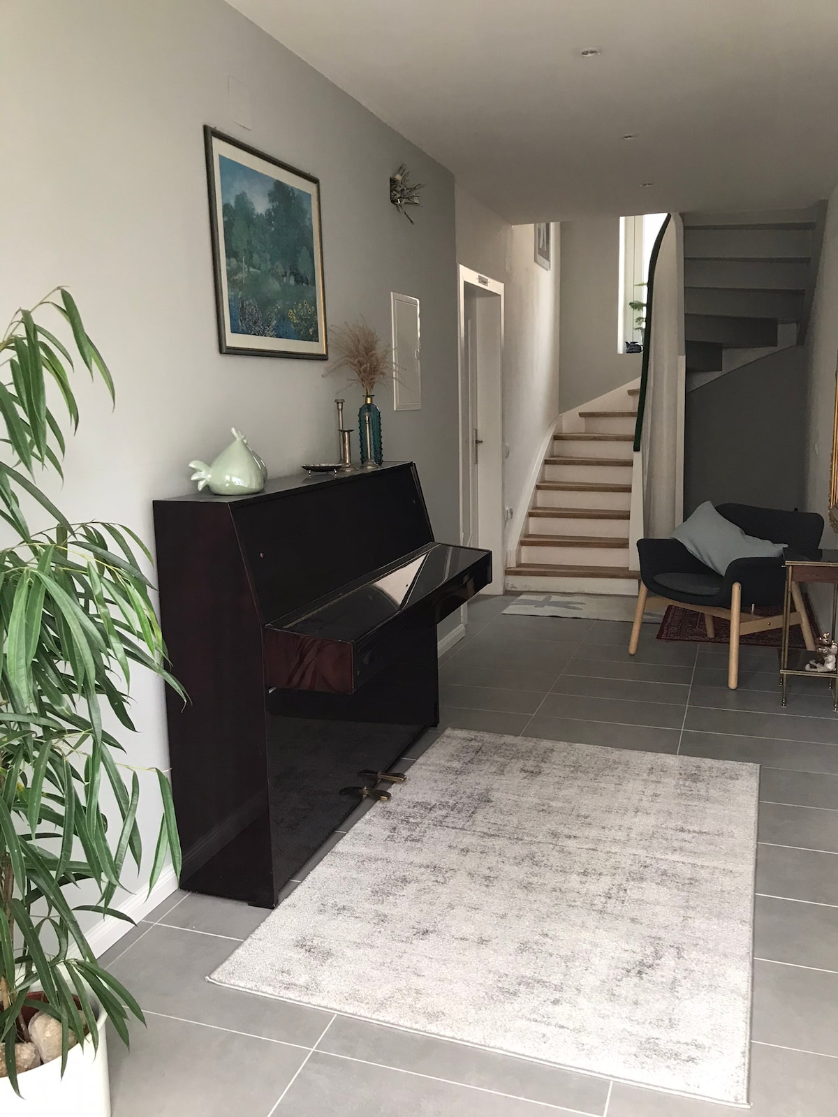 An entryway features a dark wood console table with a mirrored top, complemented by a decorative vase. A light gray area rug lies on the tiled floor, and a green plant adds a touch of nature. A staircase with wooden steps leads to the upper level.