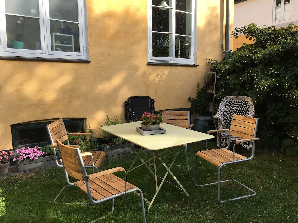 A small outdoor seating area features a light yellow table surrounded by four wooden chairs. A planter with blooming flowers sits at the center of the table. The backdrop consists of a vibrant yellow wall and lush greenery, enhancing the space's inviting feel.