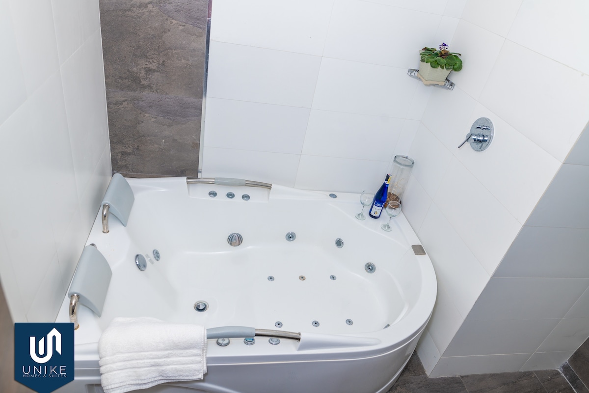 A cozy spa bath is showcased with a smooth curved design and multiple jets, set against crisp white tiles. A small plant adorns the wall shelf, while a bottle and glass sit nearby, offering a sense of relaxation.